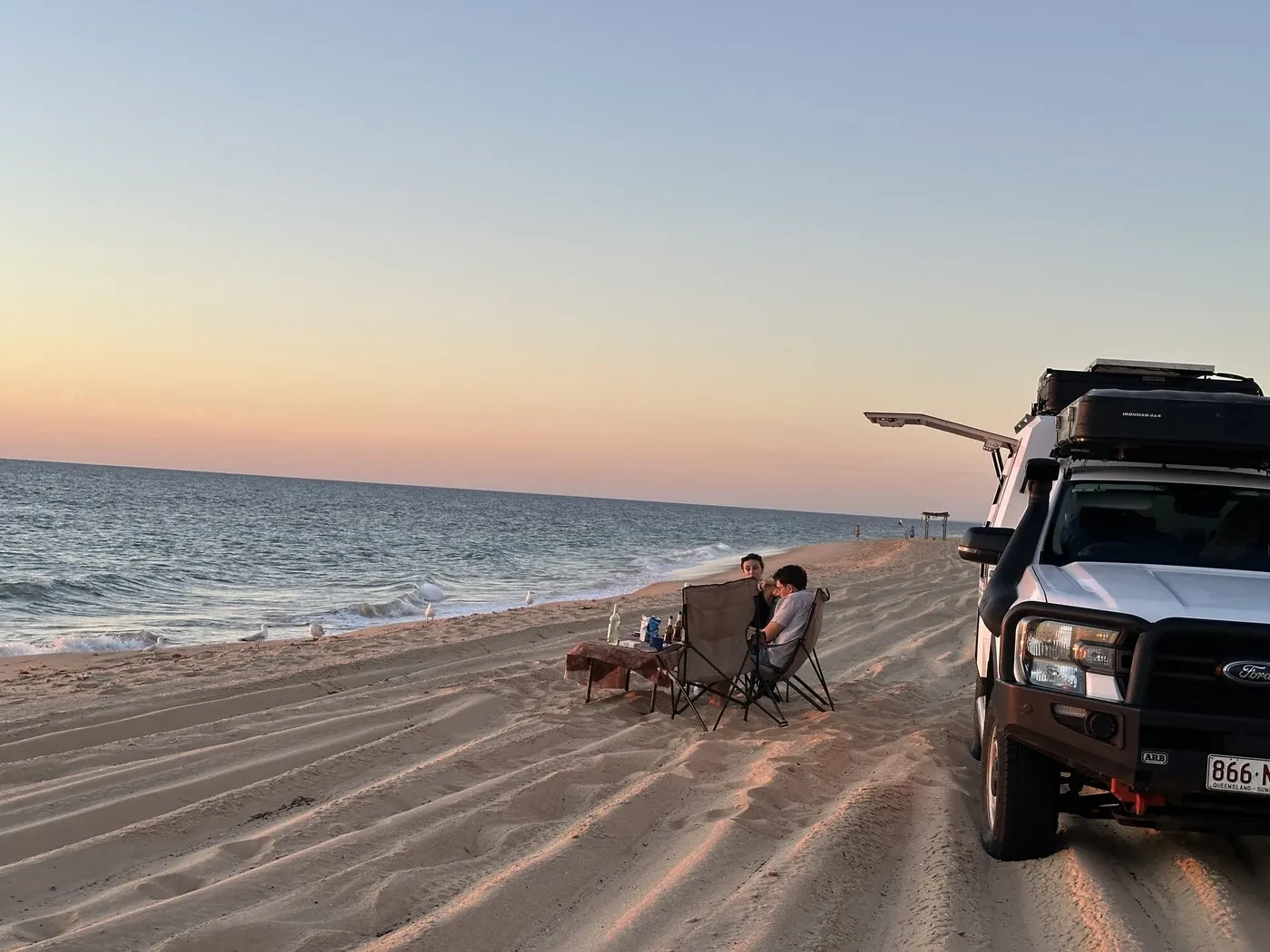 Two travellers enjoying sunset dinner in camp chairs on a golden sand beach next to their Britz 4WD Warrior with seagulls nearby