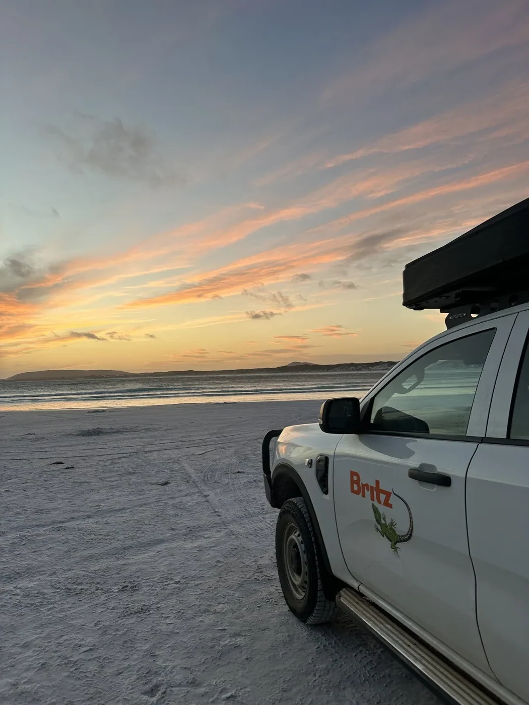 Britz 4WD Warrior parked on Lucky Bay beach at sunset with pink and orange sky reflected on wet sand