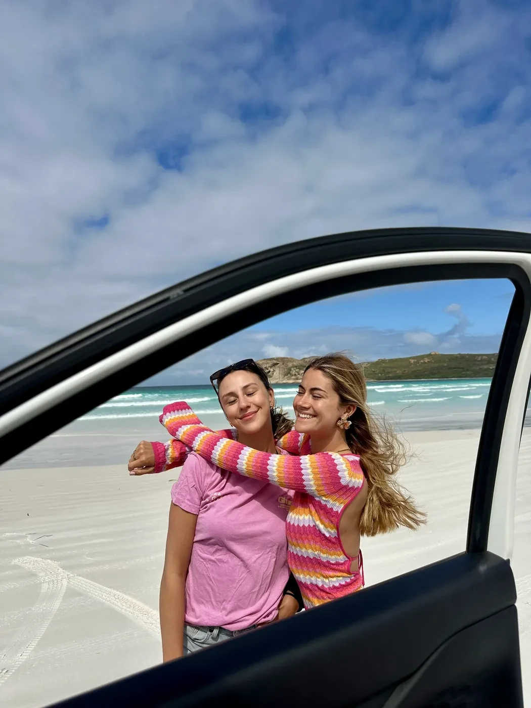 Two friends hugging and laughing through the open door of a Britz 4WD on a white sand beach with turquoise water behind them