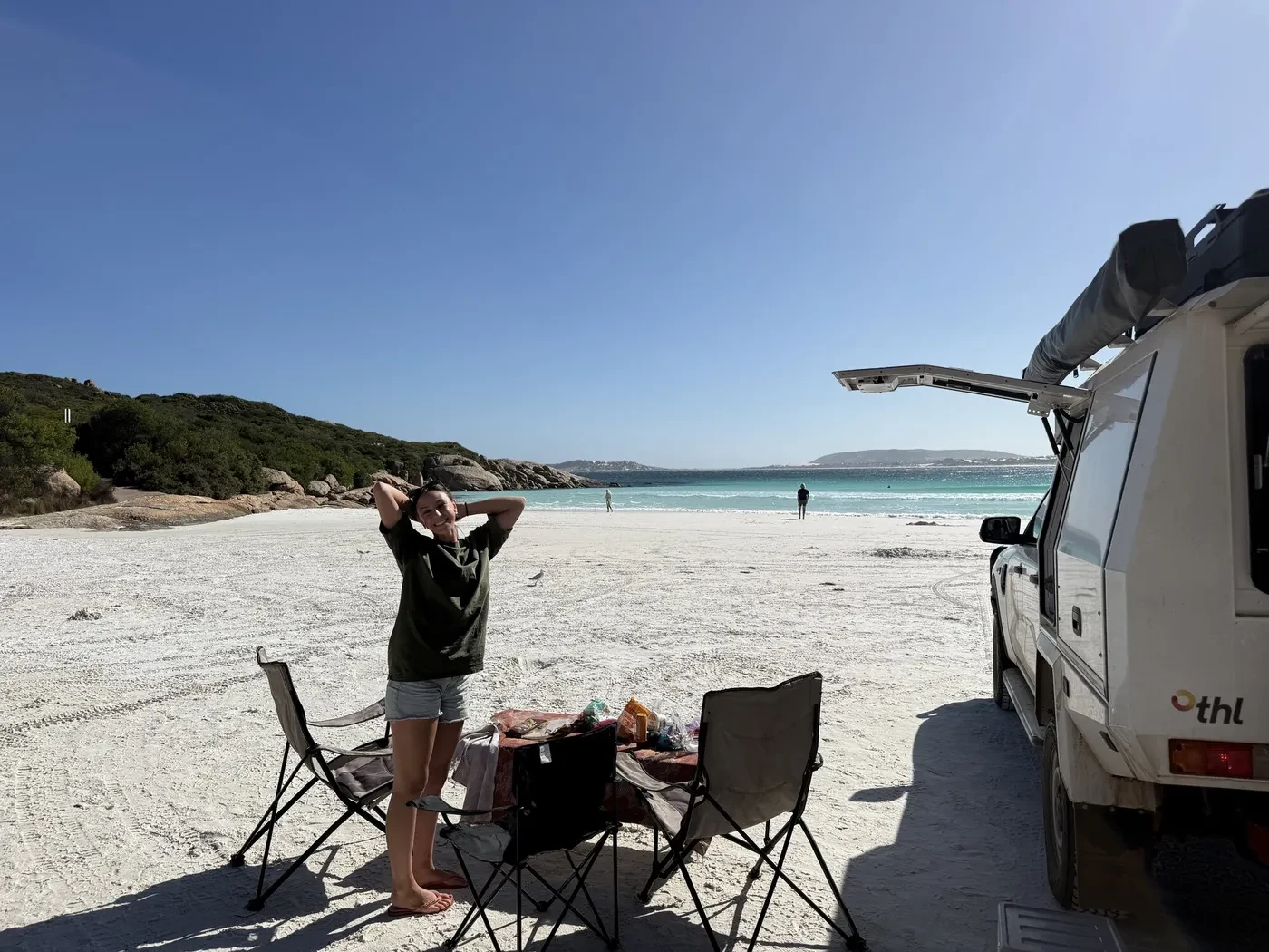 Happy traveller standing beside the Britz 4WD Warrior at Lucky Bay with white sand and turquoise water stretching into the distance