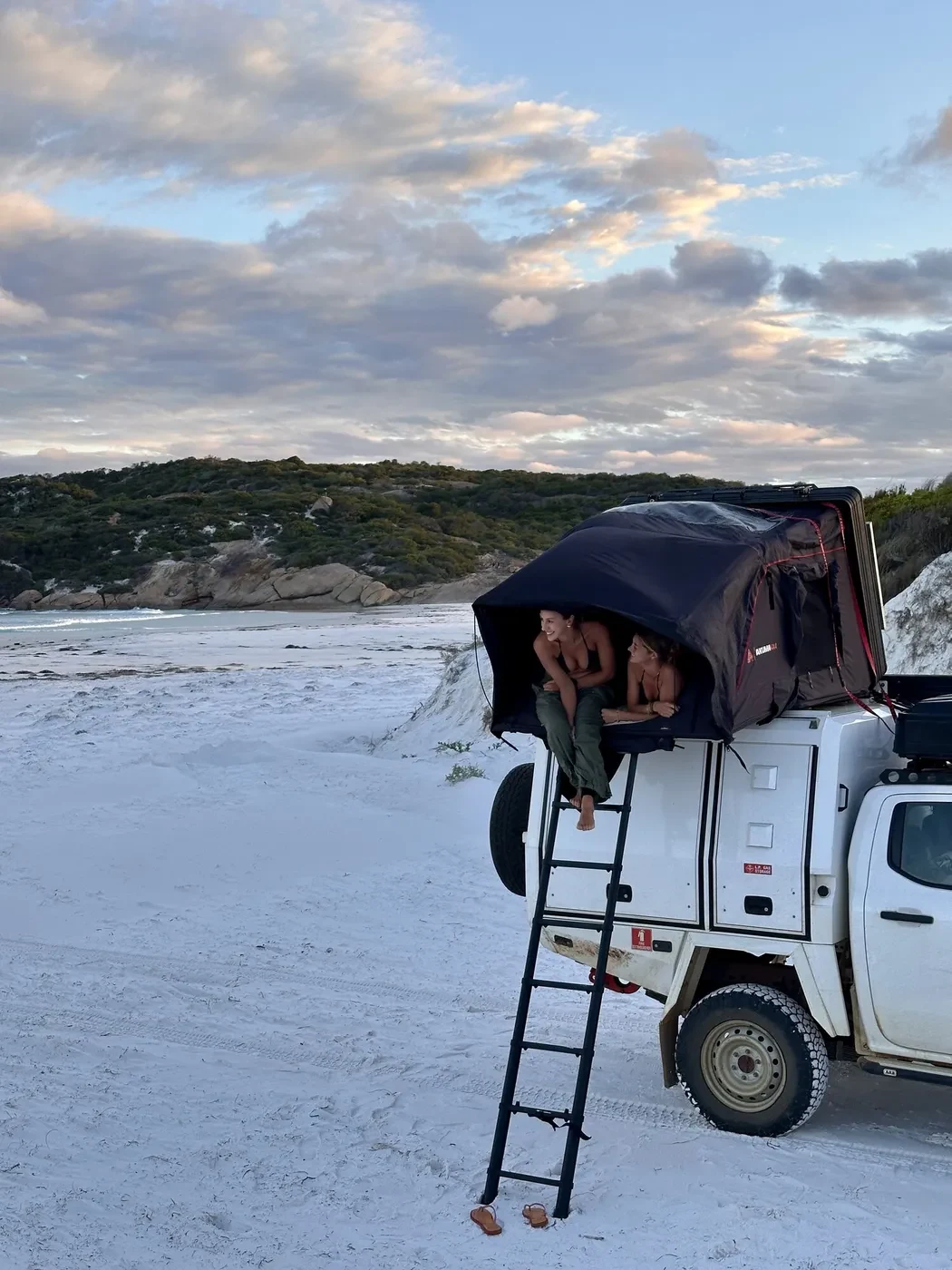 Two travellers in a Britz 4WD Warrior rooftop tent on a white sand beach at Lucky Bay with bushland and ocean in the background at sunset