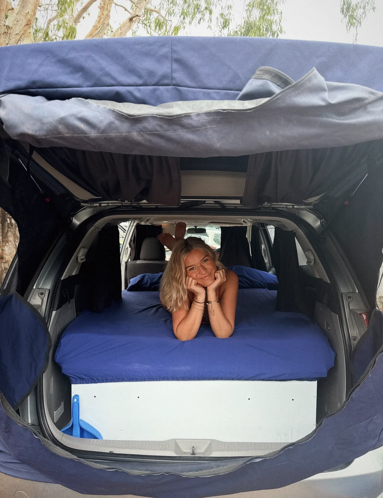 Sierra lying on the blue mattress in the back of a Spaceship Beta campervan with the rear hatch open, smiling at the camera