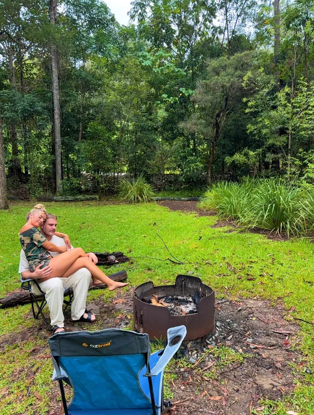 Couple sitting together by a campfire ring in a bush campground surrounded by green trees, with an OzTrail camp chair in the foreground