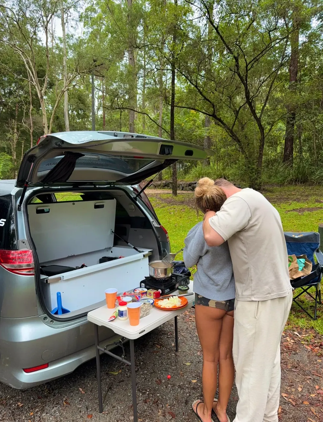 Couple cooking dinner at the slide-out camp kitchen in the rear of a Spaceship Beta campervan at a bush campsite with tall eucalyptus trees