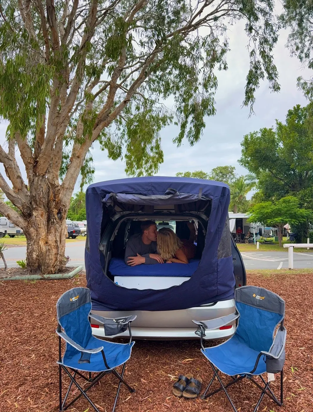 Couple kissing inside the pop-up tent of a Spaceship Beta campervan at a caravan park with eucalyptus trees and camp chairs visible