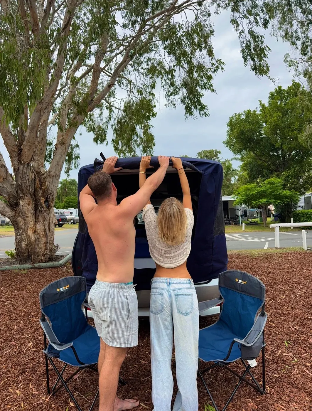Couple setting up the pop-up tent on a Spaceship Beta campervan at a caravan park with eucalyptus trees and other campervans in the background