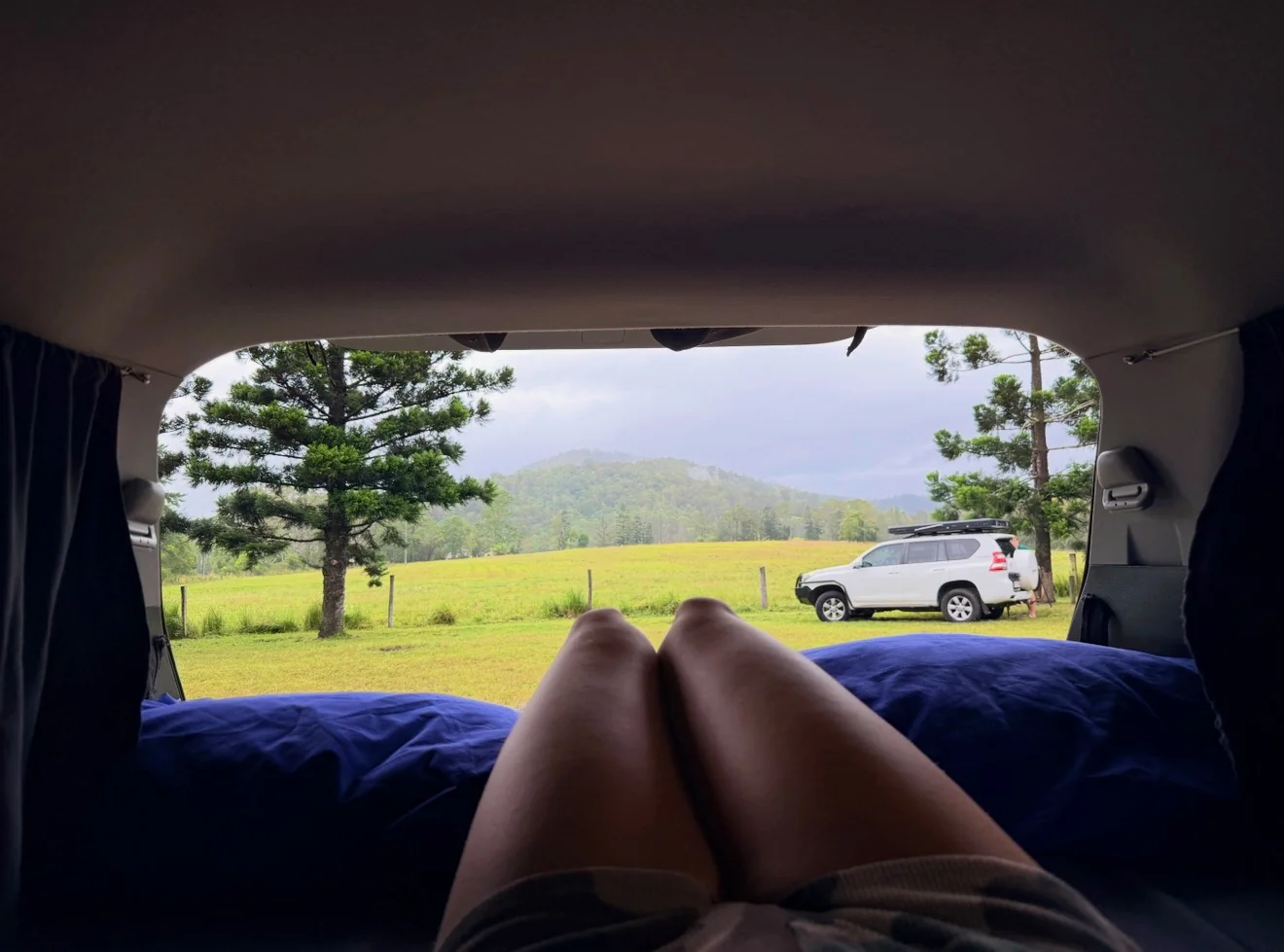 View from inside a Spaceship Beta campervan bed looking out at a green paddock with misty mountains in the background, feet visible on the blue mattress