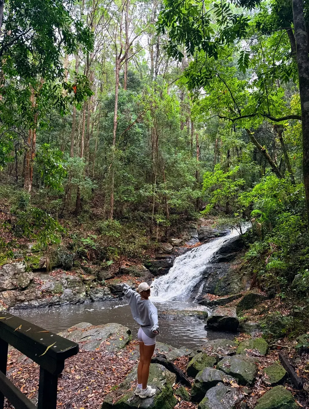 Sierra standing on rocks at the base of a waterfall in the Sunshine Coast hinterland rainforest, wearing a grey hoodie and cap